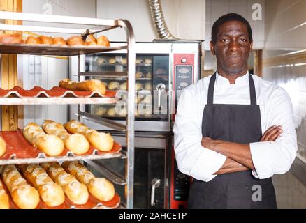 Zuversichtlich Baker in schwarze Schürze mit Waffen in der Nähe von professionellen Brotbackofen in kleine Bäckerei gekreuzt Stockfoto
