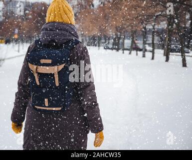 Glückliche junge europäische Frau in einem gelben Strickmütze und Fäustlinge mit einem Rucksack, lacht und lässt unter dem Schneefall. Ansicht von der Rückseite. Retro Effekt Stockfoto