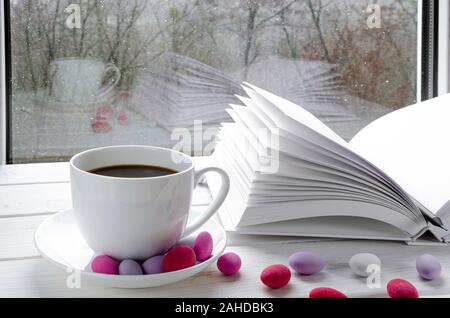 Gemütliche Konzept: Eine weiße Tasse Kaffee steht auf einem weißen Tisch neben einem weißen Buch öffnen und bunten Süßigkeiten vor dem Hintergrund eines Fensters Stockfoto