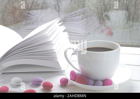 Gemütliche Konzept: Eine weiße Tasse Kaffee steht auf einem weißen Tisch neben einem weißen Buch öffnen und bunten Süßigkeiten vor dem Hintergrund eines Fensters Stockfoto