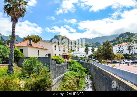 Porto da Cruz, Madeira, Portugal - Sep 24, 2019: Malerische portugiesischen Dorf, das von Hügeln und grünen Vegetation umgeben. Felsen hinter den Häusern. Strom entlang der Straße. Stockfoto