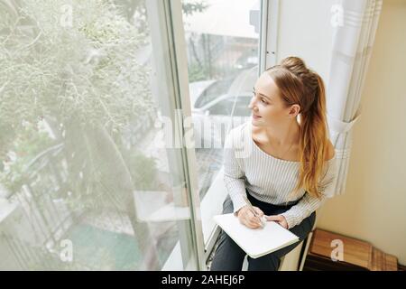 Lächelnde kreative junge Frau sitzt auf der Fensterbank und Zeichnung Baum hinter ihrem Fenster Stockfoto