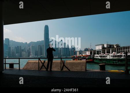 Hongkong, China - November, 2019: Silhouette eines Mannes auf Handy in Tsim Sha Tsui Star Ferry Pier und der Insel Hong Kong Skyline suchen Stockfoto