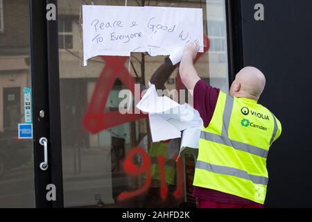 Ein Rat Reiniger entfernt ein Schild mit der Aufschrift "Frieden und guten Willen aller über anti-semitische Graffiti in Form von Nummern 9, 11 und ein Stern Davids, vor dessen Ausbau von ein Schaufenster in Belsize Park, nördlich von London. Stockfoto
