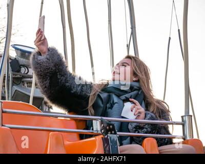 Die Princes Street Gardens, Edinburgh, Schottland, Vereinigtes Königreich. 29. Dezember 2019. Weihnachten feiern: eine hübsche blonde junge Frau Geschichten ein selfie auf ihr Mobiltelefon vor dem Star Flyer messe Runde wird ausgeschaltet Stockfoto