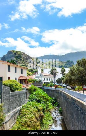 Porto da Cruz, Madeira, Portugal - Sep 24, 2019: Malerische portugiesischen Dorf umgeben von Bergen und grünen Vegetation umgeben. Felsen hinter den Häusern. Strom entlang der Straße. Stockfoto