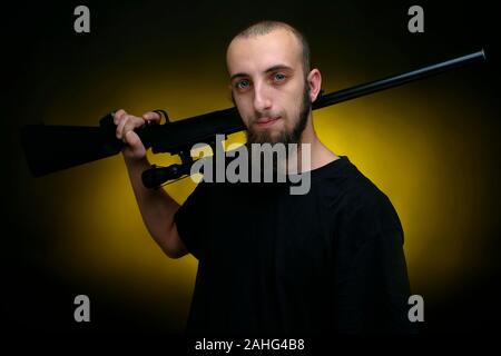 Porträt eines Mannes mit einer Schrotflinte Blättern über die Schulter. Hübscher Kerl Schuß gegen dunkel gelben Hintergrund Stockfoto