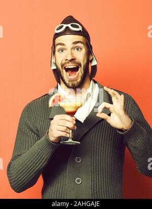 Schöne bärtige aviator Mann mit dem langen Bart auf lächelnde Gesicht holding Glas alkoholfreien Cocktail in Grau in Form gestrickter Pullover mit Hut und Brille auf Roten studio Hintergrund Stockfoto