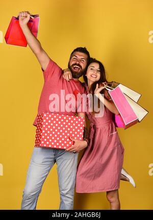 Mann mit Bart hält Red polka gestrichelten Kasten. Kerl mit Bart und Mädchen mit aufgeregten Gesichter einkaufen. Geld und Shopping Konzept. Paar in der Liebe hält Einkaufstaschen auf gelben Hintergrund. Stockfoto