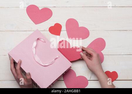 Liebe und Urlaub Konzept. Weibliche Hand, die eine rosa Herz. Rosa Papier Beutel mit Valentines auf vintage Hintergrund. Valentines Tag Karten oder Dekorationen, Ansicht von oben. Stockfoto
