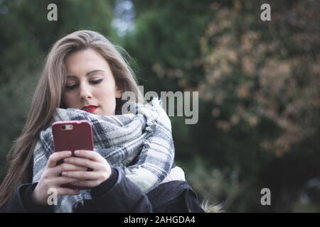 Mittellange Aufnahme der brünette Mädchen, dass smart phone mit beiden Händen im Herbst Saison Tag im Park. Junge hübsche Frau mit Handy Stockfoto