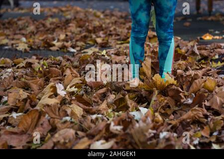 Braune Blätter auf dem Boden, die Füße von kleinen Mädchen in der Herbstsaison abdecken. Kind in grüner Leggings, das am Herbsttag im Freien steht Stockfoto