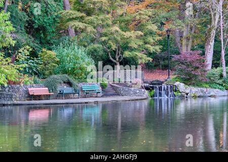 Einladende Bänke, eine Mauer aus Stein, buntes Laub und ein kleiner Wasserfall sitzen am Rande des Goodacre See im Beacon Hill Park, Victoria, British Columb Stockfoto