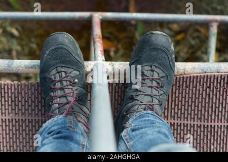 Aus dem hohen Winkel, von oben, von den dunklen Wanderschuhen eines Mannes, die auf einer Brücke stehen Stockfoto