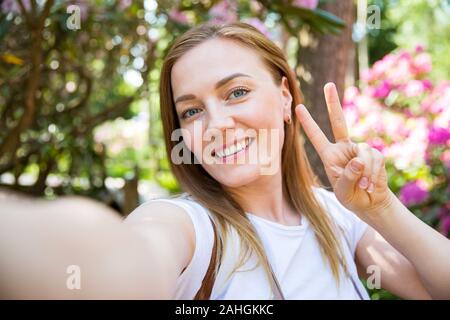 Schöne Frau, die im tropischen Wald mit Blumen unterwegs ist, auf dem Holzweg spaziert, selfie-fotos gemacht hat und glücklich lächelt. Friedenszeichen wird angezeigt Stockfoto