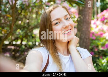 Schöne Frau, die im tropischen Wald mit Blumen unterwegs ist, auf dem Holzweg spaziert, selfie-fotos gemacht hat und glücklich lächelt. Friedenszeichen wird angezeigt Stockfoto