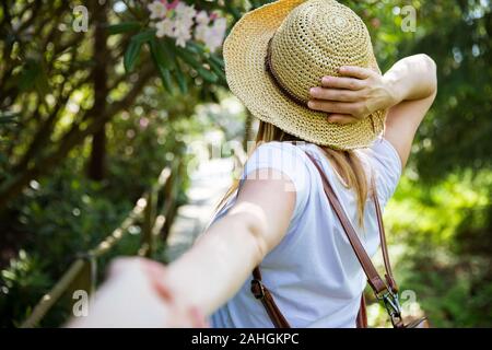 Schöne Frau im Strohhut reist im tropischen Wald, geht auf dem Holzweg und führt ihren Partner an. Tourist mit Rucksack. Folgen Sie mir Konzept. Stockfoto