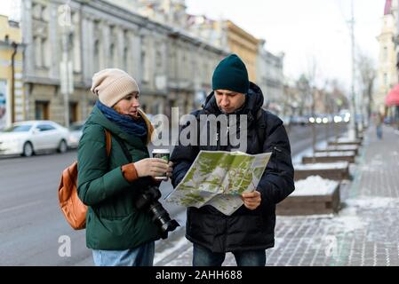 Ein paar Touristen Mädchen im Winter Kleidung mit Kamera und Tasse Kaffee und mann im winter Kleidung, im historischen Zentrum der Stadt liest ein Stockfoto