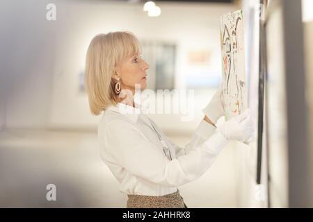 Seitenansicht Portrait von eleganten reife Frau hängenden Gemälde während der Arbeit in der modernen Kunst Galerie oder einem Museum, kopieren Raum Stockfoto