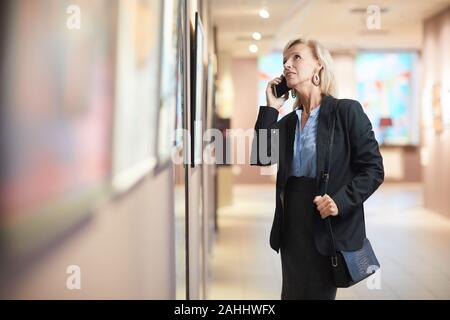 Portrait von eleganten reife Frau spricht von Smartphone und genießen Sie die Ausstellung in der Galerie oder einem Museum, kopieren Raum Stockfoto