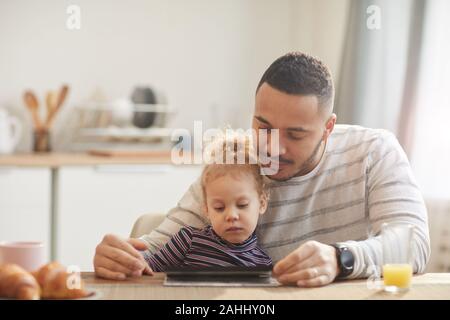 In warmen Tönen Porträt des fürsorglichen Vaters mit niedlichen kleinen Mädchen mit digitalen Tablet zusammen beim Sitzen an den hölzernen Tisch in der gemütlichen Küche, Kopie Raum Stockfoto