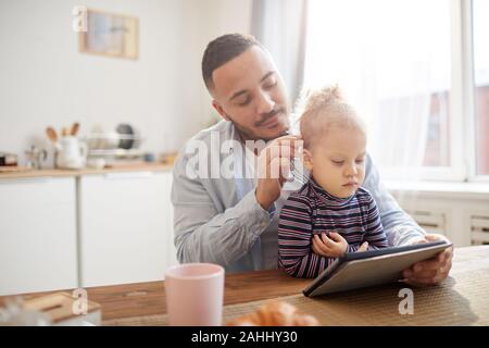 In warmen Tönen Porträt des fürsorglichen Vaters mit niedlichen kleinen Mädchen mit digitalen Tablet zusammen beim Sitzen an den hölzernen Tisch in der gemütlichen Küche Innenraum, Kopie Raum Stockfoto