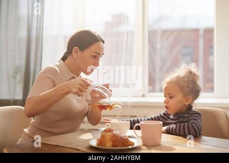 Seitenansicht Porträt des modernen Mutter gießen schwarzer Tee für süße kleine Mädchen beim Frühstück zu Hause sitzen gegen Fenster im Sonnenlicht Stockfoto