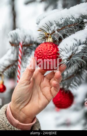 Weihnachten im Hintergrund mit Fir Tree Branches, Dekorationen und verschwommen Lichter. Weibliche Hand hängt, eine rote Kugel auf dem Weihnachtsbaum Stockfoto