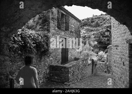 Backstreet (Rue du Porche) mit Bogen und Fußgängerin im alten Katharerdorf Minerve, Hérault, Occitanie, Frankreich. Schwarzweiß-Version. MODELL VERÖFFENTLICHT Stockfoto