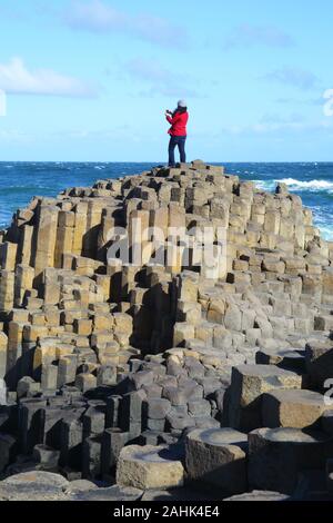 Massive Basaltsäulen Giant's Causeway mit Frau in der roten Jacke, die auf dem Gipfel Fotografieren mit Ihrem Mobiltelefon, blauer Himmel und Stockfoto