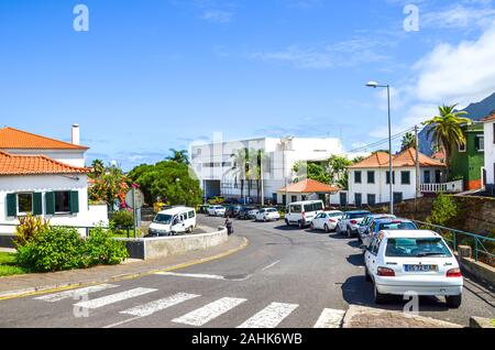 Porto da Cruz, Madeira, Portugal - Sep 24, 2019: Straße der malerischen portugiesischen Dorf. Häuser, Menschen auf der Straße, geparkte Autos. Sonnigen Tag, blauer Himmel. Zebrastreifen im Vordergrund. Stockfoto