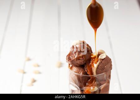 Eine Soße ist auf Schokolade Eis in einem eisbecher Glas auf einem weißen plank Tischplatte gegossen Stockfoto
