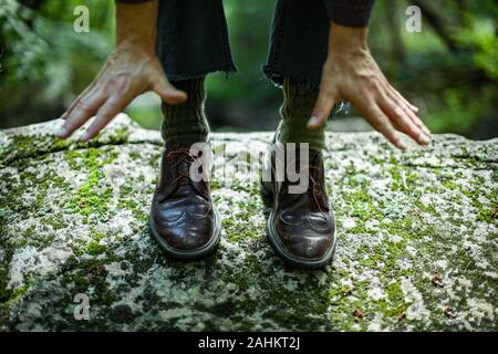 Junge männliche tragen braune Lederschuhe mit grünen hippie Wollsocken und blaue zerrissene Jeans. Nahaufnahme der beiden Hände zusammen erreichen Sie zu Boden. Stockfoto