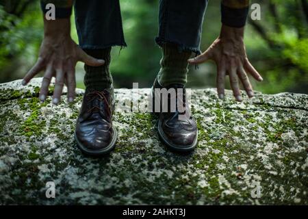 Junge männliche tragen braune Lederschuhe mit grünen hippie Wollsocken und blaue zerrissene Jeans. Nahaufnahme der beiden Hände zusammen erreichen Sie zu Boden. Stockfoto