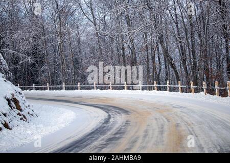 Eine geschwungene Landstraße auf der Seite eines Hügels ist im Winter gesehen, bei Eis und Schnee auf den Spuren von früheren Fahrzeugen abgedeckt. Stockfoto