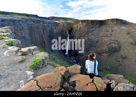 Junge Frau sitzt an den Felsen vor der spektakulären Blick auf den Wasserfall Litlanesfoss mit senkrechten Basaltsäulen umgeben, Fljotsdalshreppur m Stockfoto