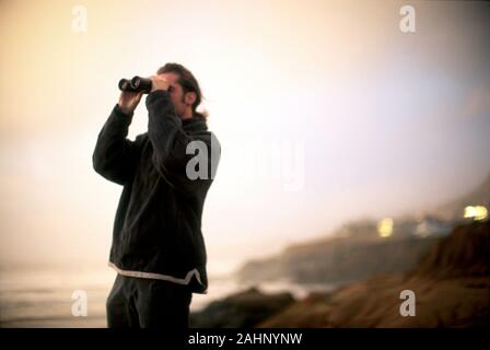 Seitenansicht eines Mannes, der durch ein Fernglas auf einer friedlichen Küste blickt. Stockfoto