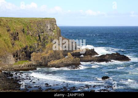 Dramatische basalt Küste mit Wellen auf die Felsen brechen und ein blauer Himmel mit weißen Wolken Puffy, Antrim Coast, County Antrim, Nordirland, Großbritannien. Stockfoto