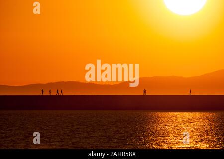 Silhouetted Wanderer auf einem Steg im Moment von einem goldenen Sonnenuntergang mit Blick auf Meer und Berge im Hintergrund Stockfoto