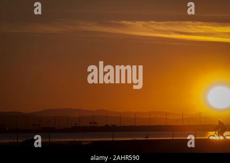 Silhouetted Radfahrer auf einem Deich im Moment von einem goldenen Sonnenuntergang mit Blick auf Meer und Berge im Hintergrund Stockfoto