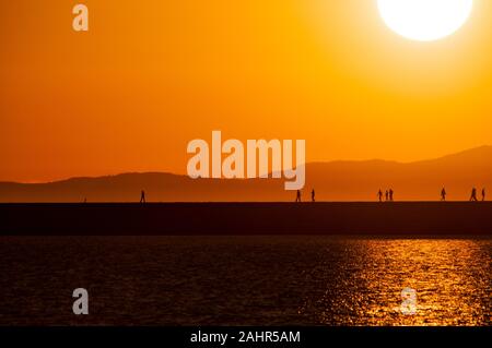 Silhouetted Wanderer auf einem Steg im Moment von einem goldenen Sonnenuntergang mit Blick auf Meer und Berge im Hintergrund Stockfoto