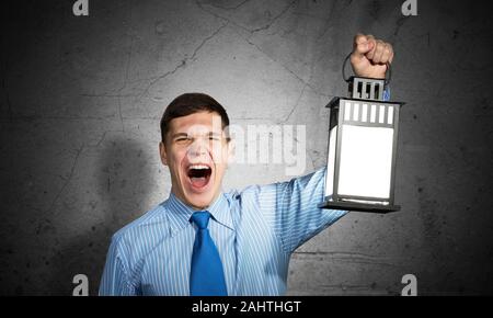 Schreien Geschäftsmann Holding leuchtende Laterne auf Hintergrund grunge Wand. Vorderansicht der junge Mensch in Hemd und Krawatte auf der Suche nach Etwas in d Stockfoto