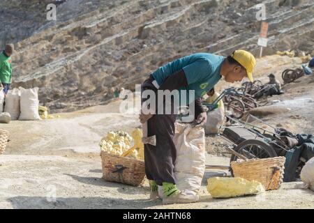 Ein Indonesien Schwefel Miner bricht seine Last von Schwefel in kleinere Stücke, bevor er transportiert es die Ijen Vulkan, Ost Java, Indonesien. 08/12/19. Stockfoto