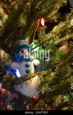 Hausgemachte gestrickte Schneemann in einen Hut und Schal auf die Zweige von einem Weihnachtsbaum, mit einer bunten Girlande geschmückt. Stockfoto