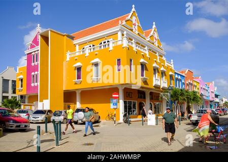 Eine Reihe von hell gemalten Geschäften und Gebäuden in einer Straße in Kralendijk, Bonaire in der Karibik. Touristen und Anwohner mischen sich in der belebten Straße Stockfoto