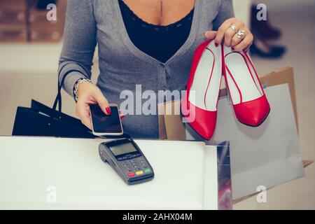 Zugeschnittenes Bild einer Frau Schuhe im Shop Kasse kauft, ist Sie bezahlen mit Kontaktlosen smartphone Zahlung Stockfoto