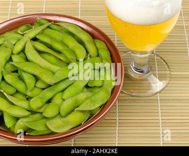 Gekochte edamame in einer Schüssel mit einem Glas Bier auf Bambus Matte. Grüne Sojabohnen, maodou, in Salzwasser gekocht. Beilage und leckeren Snack. Sojabohnen. Stockfoto