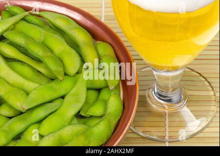 Gekochte edamame in brauner Schale mit Glas Bier auf Bambus Matte. Grüne Sojabohnen, maodou, in Salzwasser gekocht. Beilage und leckeren Snack. Sojabohnen. Stockfoto