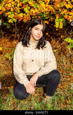 Junge hübsche stilvoll gekleideten Mädchen mit beige Pulli und schwarze Jeans sitzt in den bunten Herbst Wald im Oktober in Bayern, Deutschland Stockfoto