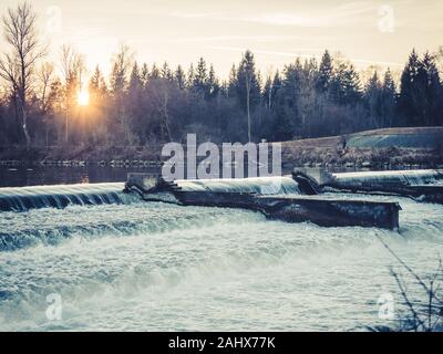 Wasser über ein Wehr bei Sonnenuntergang mit Sonnenstrahlen im Winter fliesst, Augsburg Stockfoto